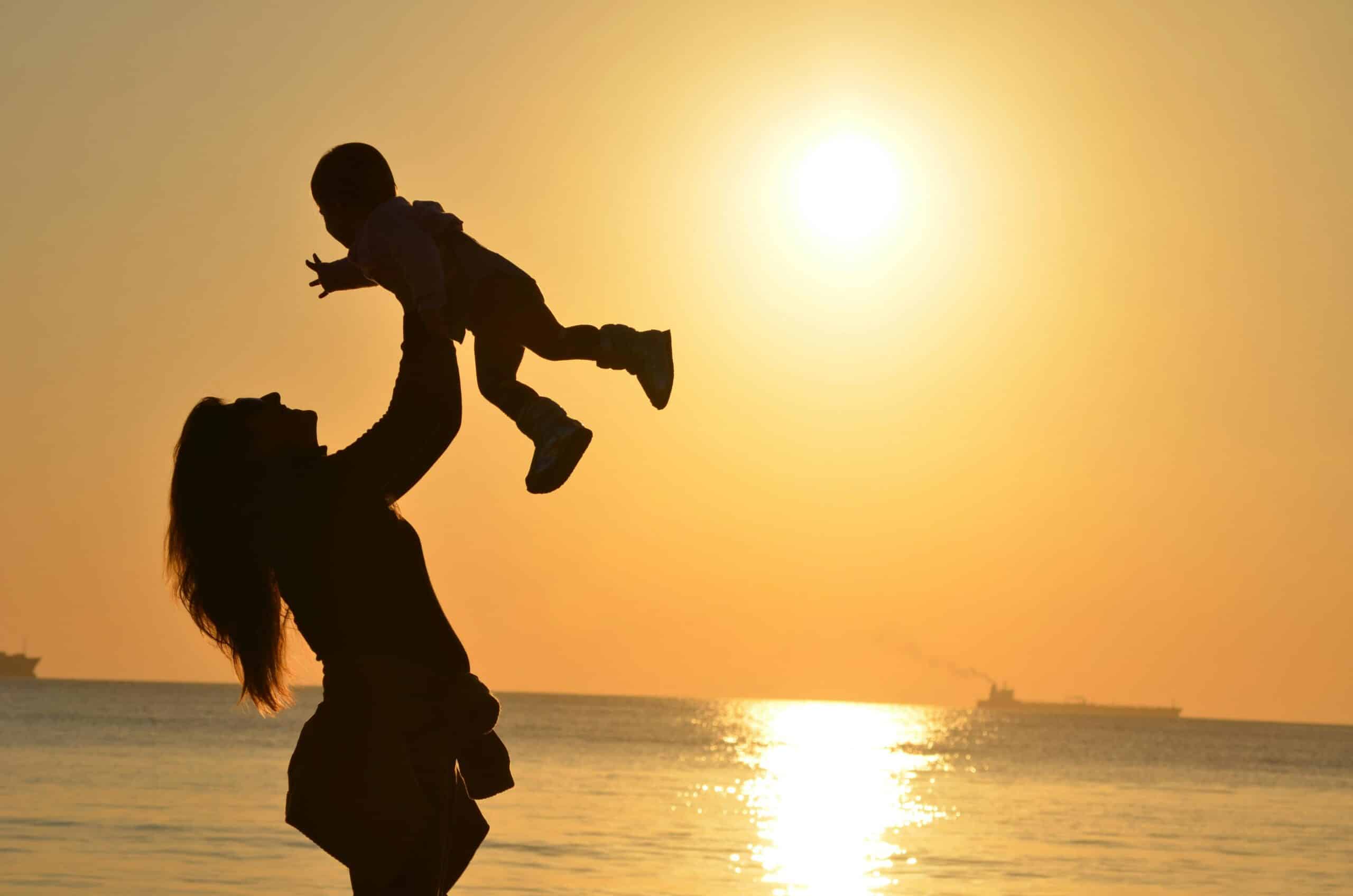 A mother holds her child high in the air whilst the sun sets in the background.