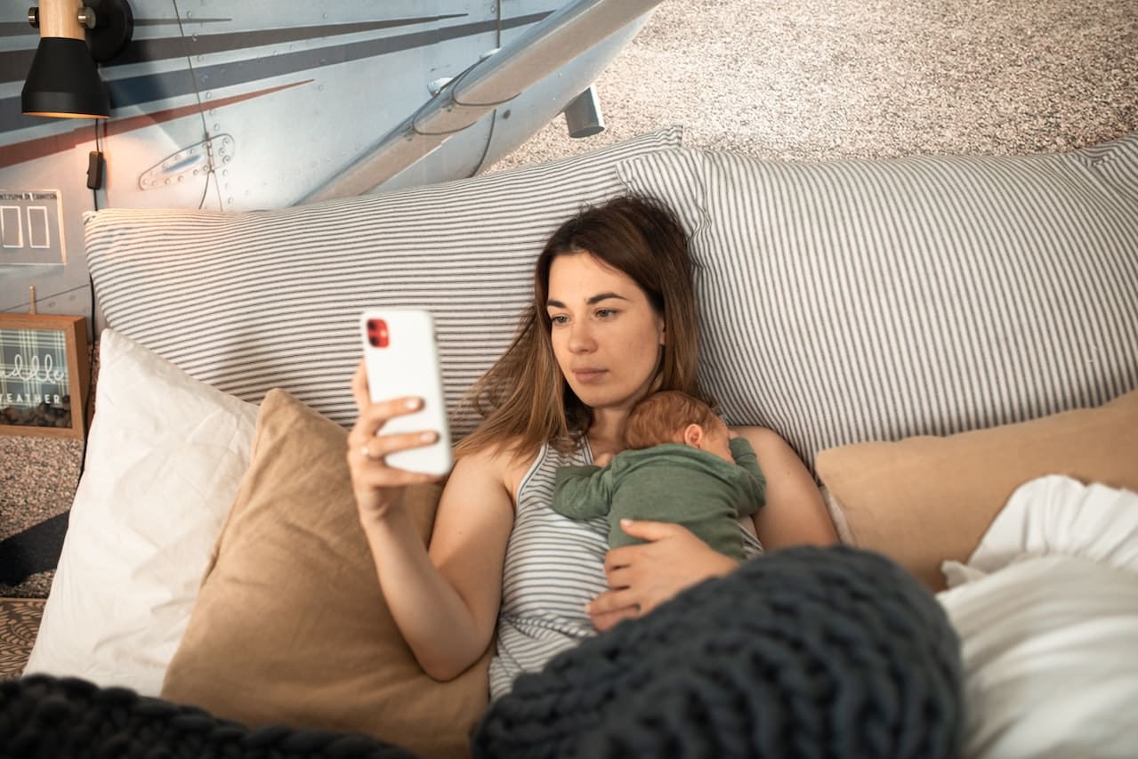 A mum lying on a bed with her baby on her chest while on her mobile phone.