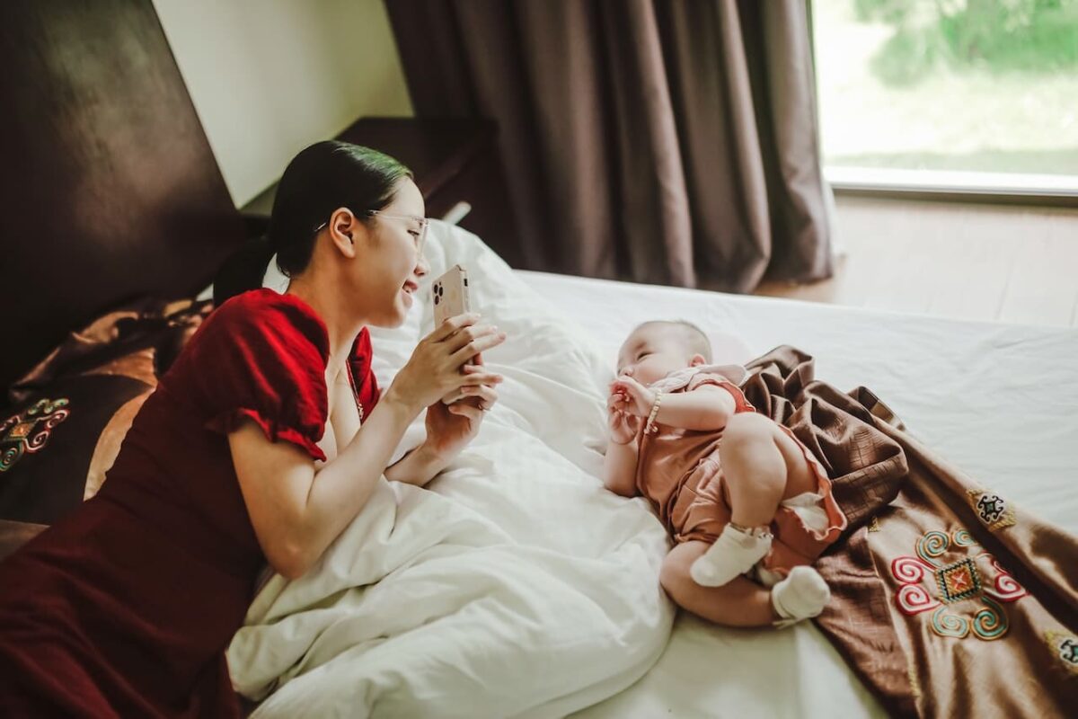 Mother and baby lying on bed while mother shows baby smartphone.